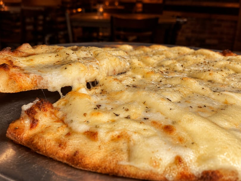 A close-up of a cheesy white pizza with herbs, a slice being lifted, showing melted cheese.
