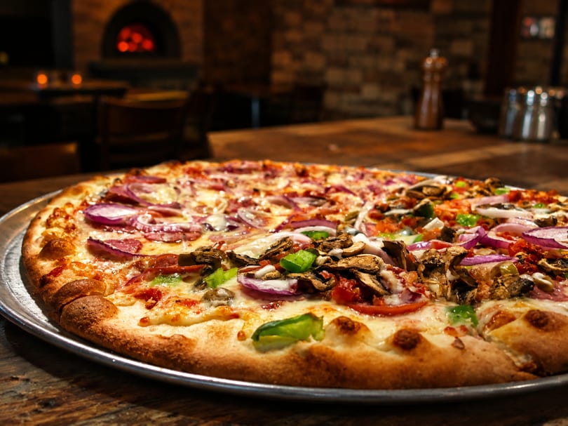 A large, half-and-half pizza with various toppings sits on a table with a brick oven in the background.